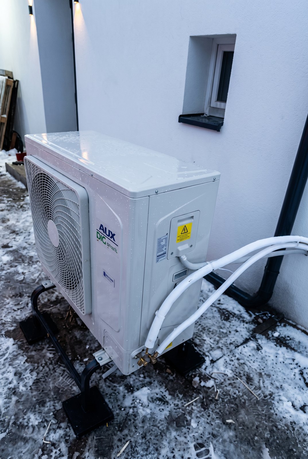 Jednostka zewnętrzna pompy ciepła AUX 12 kW przy ścianie domu zimą, śnieg